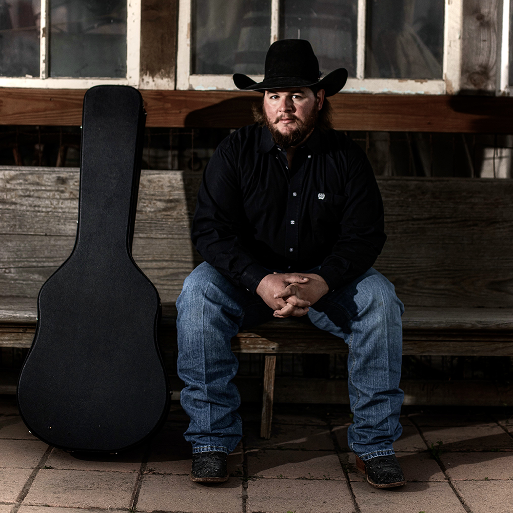 Ryan Talley, Texas country music singer-songwriter, sitting on a rustic bench with guitar case. Professional country artist for hire. Book Ryan Talley Music for live shows, weddings, and events